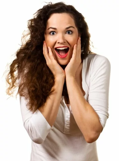 A woman with long curly hair in a white shirt holds her face with both hands, displaying a surprised or excited expression—just like receiving great news from a successful email marketing campaign—against a plain white background. stage-one-labs A woman with long curly hair in a white shirt holds her face with both hands, displaying a surprised or excited expression—just like receiving great news from a successful email marketing campaign—against a plain white background. stage-one-labs